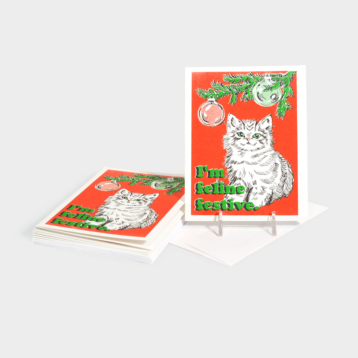 Greeting cards with an illustrated kitten under a pine tree branch with hanging ornaments. 