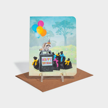 Greeting card with illustration of a raccoon standing atop a pile of garbage, wearing a party hat and holding balloons.